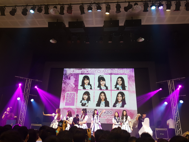 Eine Gruppe von Menschen performs auf einer hell beleuchteten Bühne mit Trägern, während eine Menge darunter Fotos macht; ein großer Bildschirm im Hintergrund zeigt Bilder der K-Pop-Gruppe BK48.