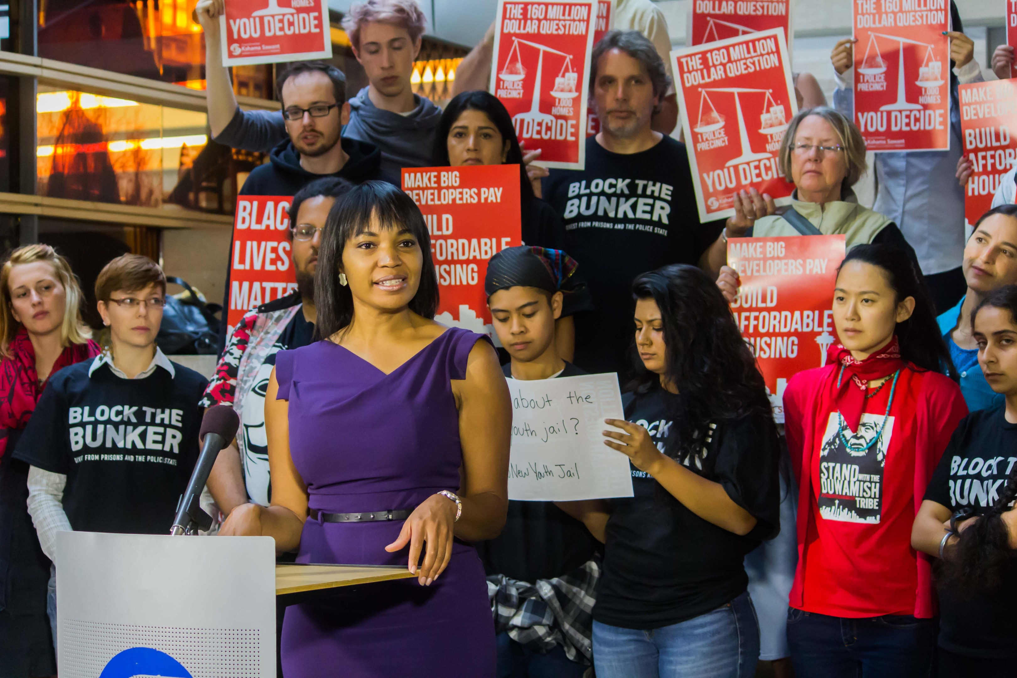Eine Black-Lives-Matter-Aktivistin spricht an einem Rednerpult mit Mikrofon zu einer Menge mit Protestschildern, im Hintergrund Scheinwerfer.