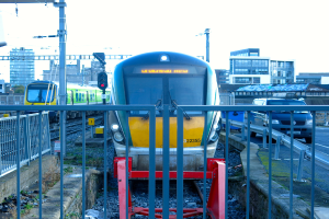 Zwei Züge auf der Schiene mit einem Zaun im Vordergrund, Pfosten auf dem Land, einer mit einer Signalleuchte, ein Auto auf der Straße rechts, Gebäude im Hintergrund und der Himmel oben.