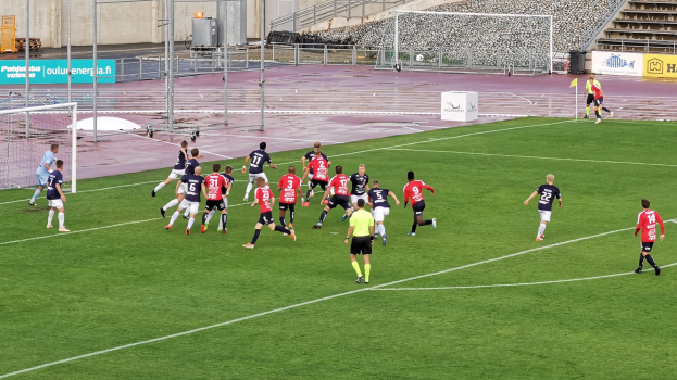 Gruppe von Menschen, die Fußball auf einem Feld spielen, umgeben von Zuschauern in Stadionrängen, umgeben von Pfosten, Tafeln mit Text, Geländern, Stufen und anderen Gegenständen.