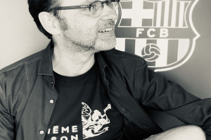 Ein Mann mit Brille und schwarzem Hemd sitzt lächelnd vor einem schwarz-weißen FC Barcelona-Logo.