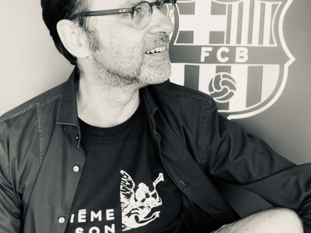 Ein Mann mit Brille und schwarzem Hemd sitzt lächelnd vor einem schwarz-weißen FC Barcelona-Logo.