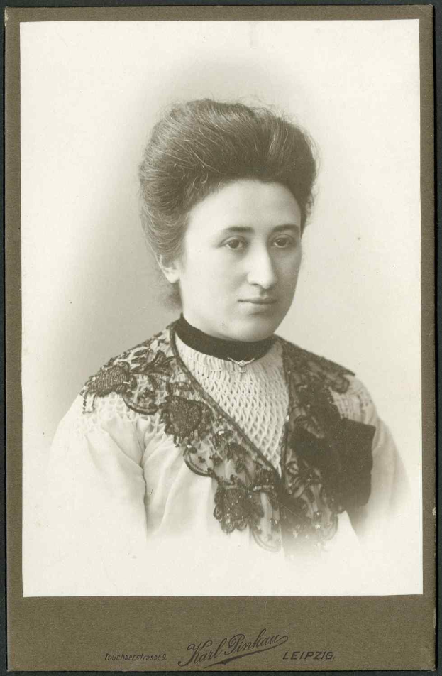 Schwarzes und weißes Foto einer Frau in einem weißen Kleid mit einem gemusterten Schultertuch, identifiziert als Karl Pinkau Leipzig, die direkt in die Kamera schaut und einen ernsten Ausdruck hat.