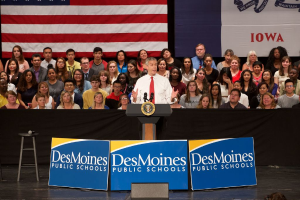 Ein Mann in einem weißen Hemd und roter Krawatte steht an einem Rednerpult mit drei Tafeln mit Text, einem Hocker mit Mikrofon und einer sitzenden Menge im Hintergrund, mit einer Flagge und zusätzlichem Text im Bild.