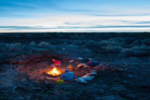 Ein Lagerfeuer mit Menschen, die darauf sitzen und liegen, mit M√ľtzen tragend, mit einem Rucksack und anderen Gegenst√§nden auf dem Boden, umgeben von B√§umen und Himmel.