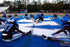 Gruppe junger Männer in Sportkleidung, Helmen und Schuhen sitzend auf einem Fußballfeld mit Bäumen, Pfählen, Gebäuden und einem klaren blauen Himmel im Hintergrund, neben einer Treppe mit Geländern.