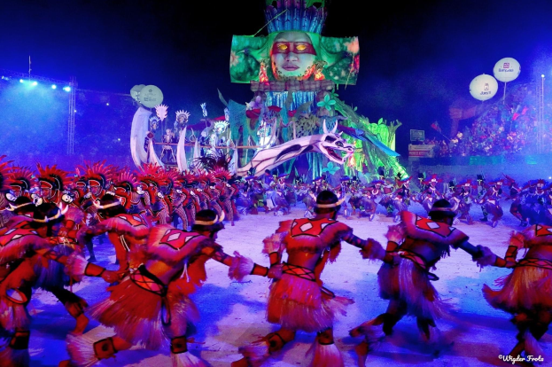 Eine Gruppe von Menschen in bunten Kostümen tanzt auf einer Bühne vor einem sitzenden Publikum, mit Lichtern, Polen, Ballons und einem Bildschirm, der eine Person im Hintergrund zeigt, unter einem sichtbaren Himmel, bei der Rio-Karneval 2014.
