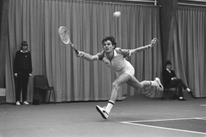 Ein Mann schwingt einen Tennis-Schläger gegen einen Ball, mit zwei stehenden und zwei sitzenden Personen im Hintergrund und einem Vorhang als Rahmen.