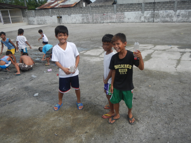 Eine Gruppe junger Jungs, die zusammen in einer Parkplatz steht, einige halten Flaschen, mit Häusern, Bäumen, Pfählen und einem klaren blauen Himmel im Hintergrund.
