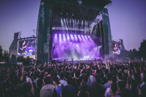 Eine große Menschenmenge auf einem Musikfestival schaut Künstlern auf einer beleuchteten Bühne mit Instrumenten und Bildschirmen zu, die vor einem Gebäude, Bäumen und einem klaren blauen Himmel aufgebaut ist.