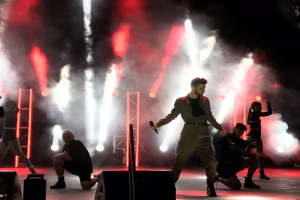 Drake, der ein Mikrofon hält, performs auf der Bühne beim 2018 Coachella Valley Music and Arts Festival, umgeben von einer Gruppe von Menschen in Hockstellung, mit Lautsprechern, einer Person mit Videokamera, Metallstangen und einer Decke mit Deckenleuchten.