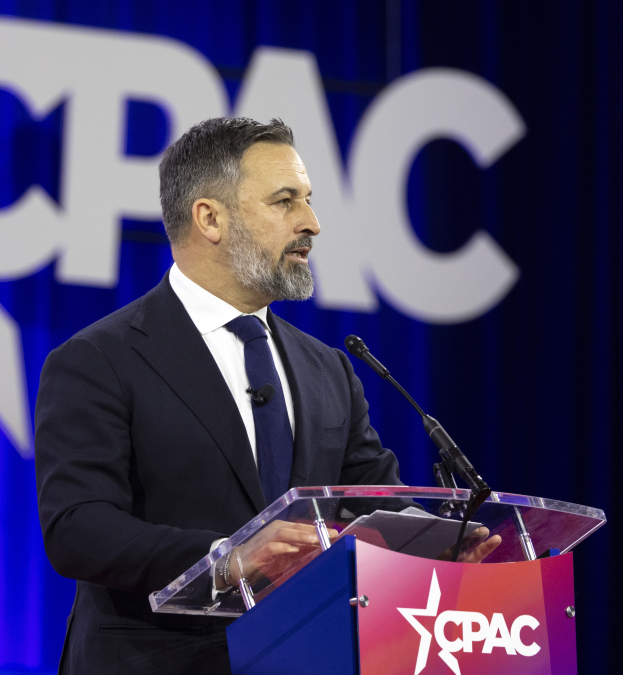 Ein Mann im Anzug und Krawatte steht an einem Podium mit zwei Mikrofonen und hält eine Rede mit einem Banner im Hintergrund.