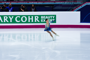 Eine Frau in einem blauen Kleid skatet auf einer Eisbahn während eines professionellen Eiskunstlauf-Events, mit einer Menge, die Kameras hält und ein Schild mit der Aufschrift 'Tessa Virtue und Scott Moir' im Hintergrund.