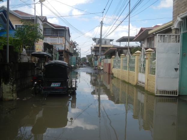 Eine überflutete Straße mit einem geparkten Motorrad, Gebäuden, Bäumen, Polen und Drähten auf beiden Seiten, unter einem bewölkten Himmel.