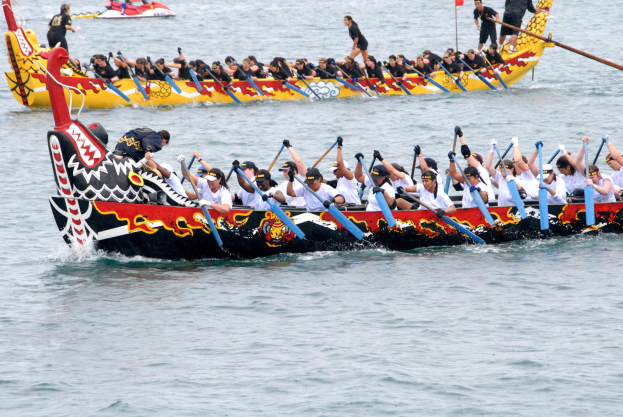 Eine Gruppe von Menschen in einem Drachenboot, das auf dem Wasser rennt und Paddel hält, mit einem anderen Boot und einer Flagge im Hintergrund.