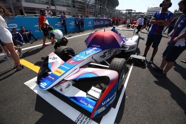 Formula E-Fahrzeug mit weißer, blauer, roter und schwarzer Lackierung auf einer Rennstrecke geparkt, umgeben von Menschen, mit einem Schirm im Vordergrund und Rennstrecken-Infrastruktur im Hintergrund unter einem klaren blauen Himmel.