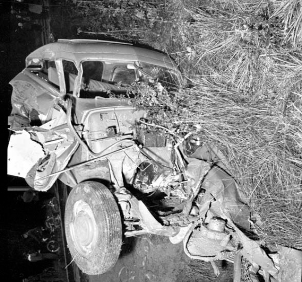 Ein Schwarz-Weiß-Foto eines schwer beschädigten Autos am Straßenrand mit einer zertrümmerten Front, zersplitterten Fenstern, einem Baum im Hintergrund und verstreuten Gegenständen.