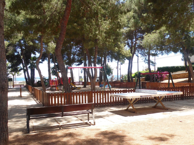 Eine Parkszene mit einem zentralen Tischtennistisch, umgeben von Bänken, einem Zaun, Schaukeln, Pfählen, Pflanzen und Bäumen, mit einem Wasserlauf und einem klaren blauen Himmel im Hintergrund.