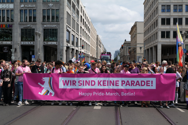 Eine Gruppe von Menschen marschiert auf einer Straße in Berlin mit einem pinken "Happy Pride March"-Schild, während Gebäude, Laternenmasten und Verkehrszeichen die Straße säumen und der Himmel bewölkt ist.