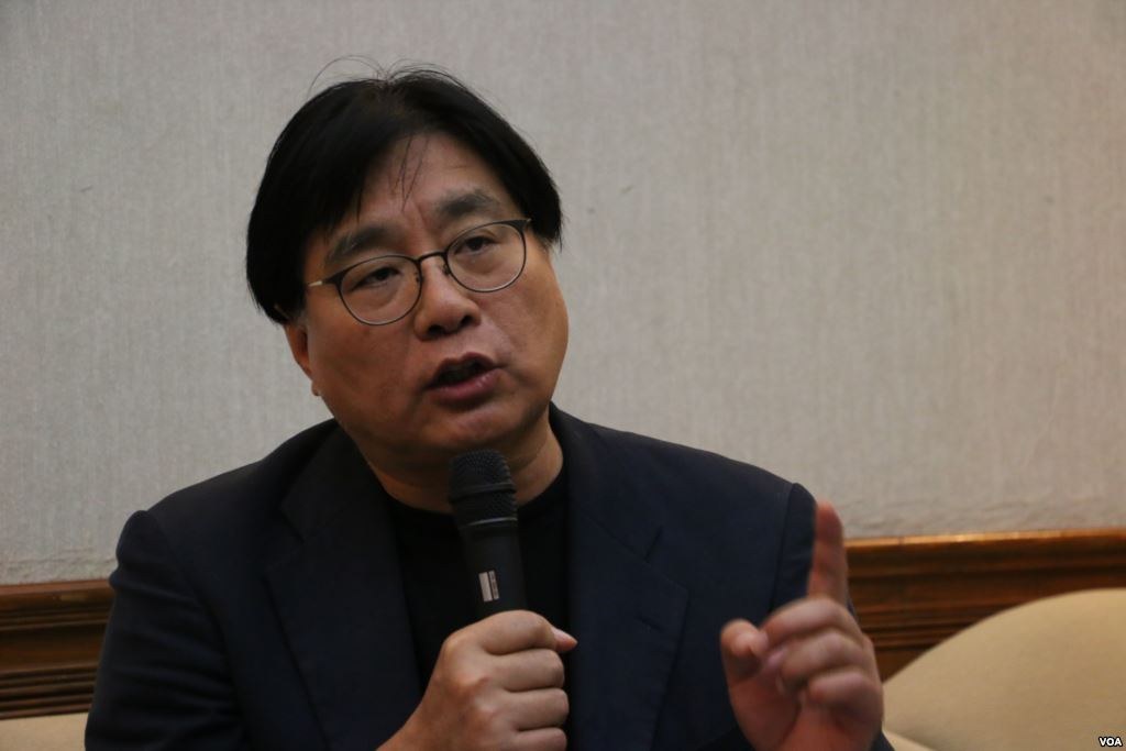 Ein Mann im Anzug und mit Brille spricht in ein Mikrofon, mit einer Wand im Hintergrund.