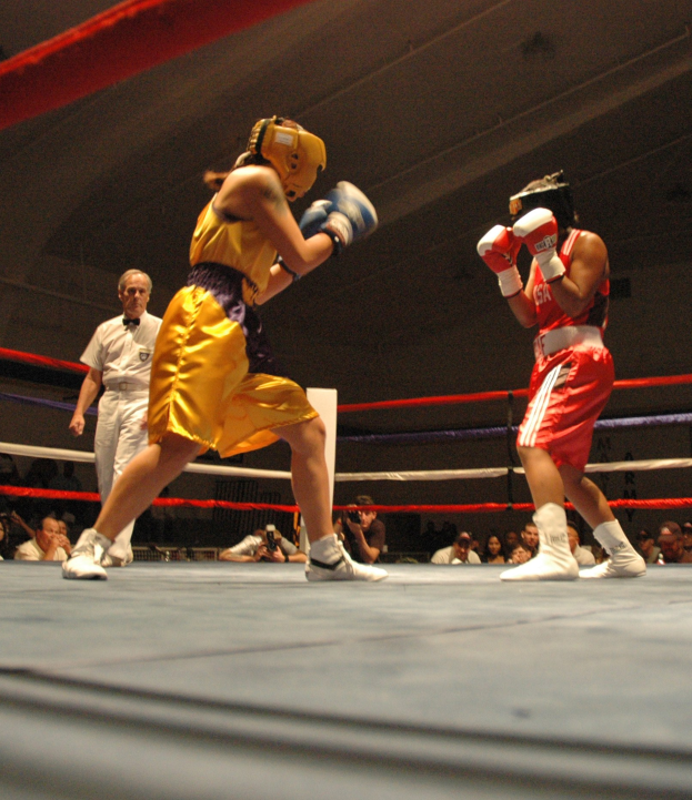 Zwei Boxer kämpfen im Ring mit einem Schiedsrichter und Zuschauern im Hintergrund, einige halten Kameras, unter einer sichtbaren Decke.