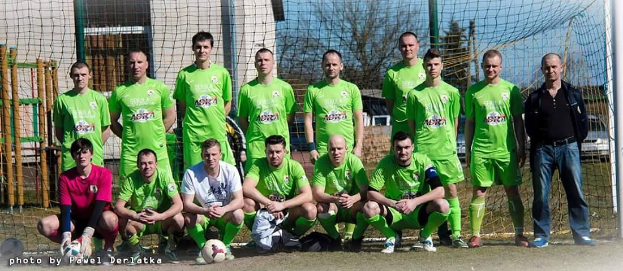 Eine Gruppe von Männern in grünen T-Shirts steht auf einem Fußballfeld, einige hocken, mit einem Ball vor ihnen und einem Torpfosten rechts daneben, vor einem Hintergrund aus Bäumen, Gebäuden und Fahrzeugen.