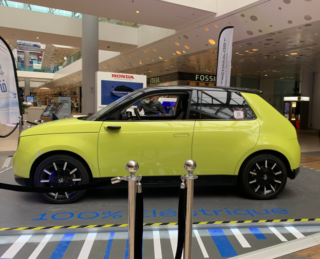 Ein gelbes Wasserstoff-elektrisches Auto steht vor einem Gebäude, umgeben von Absperrungen und Schildern, mit Menschen in der Nähe und einer Topfpflanze.