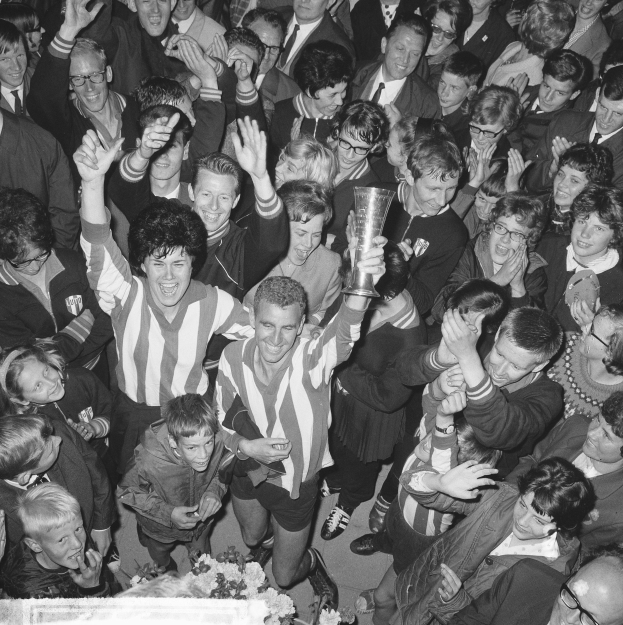 Ein Schwarz-Weiß-Bild einer Gruppe von Menschen, die mit einem Pokal feiern, der Mittelpunkt hält die Tasse, umgeben von einer Menge und Blumen unten.