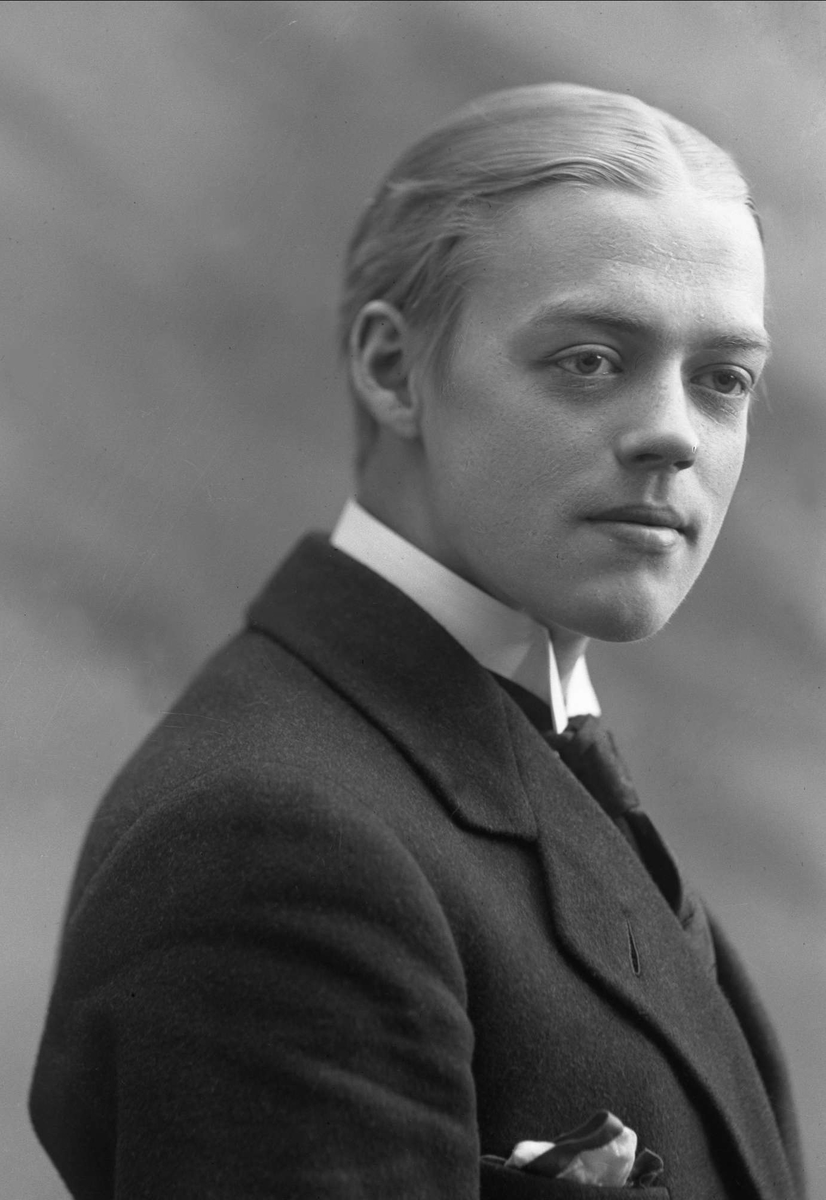 Ein Schwarz-Weiß-Foto eines Mannes im Anzug, mit unscharfem Hintergrund und Text unten, der eine weiße Krawatte und ein gestärktes weißes Hemd trägt, mit ordentlich gekämmtem Haar und einem ernsten Gesichtsausdruck.