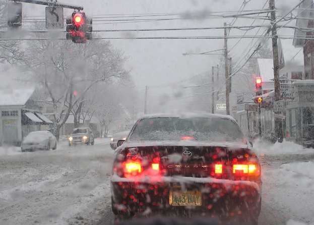 Eine ruhige, schneebedeckte Straße mit Häusern und Bäumen auf beiden Seiten und wenigen Fahrzeugen.