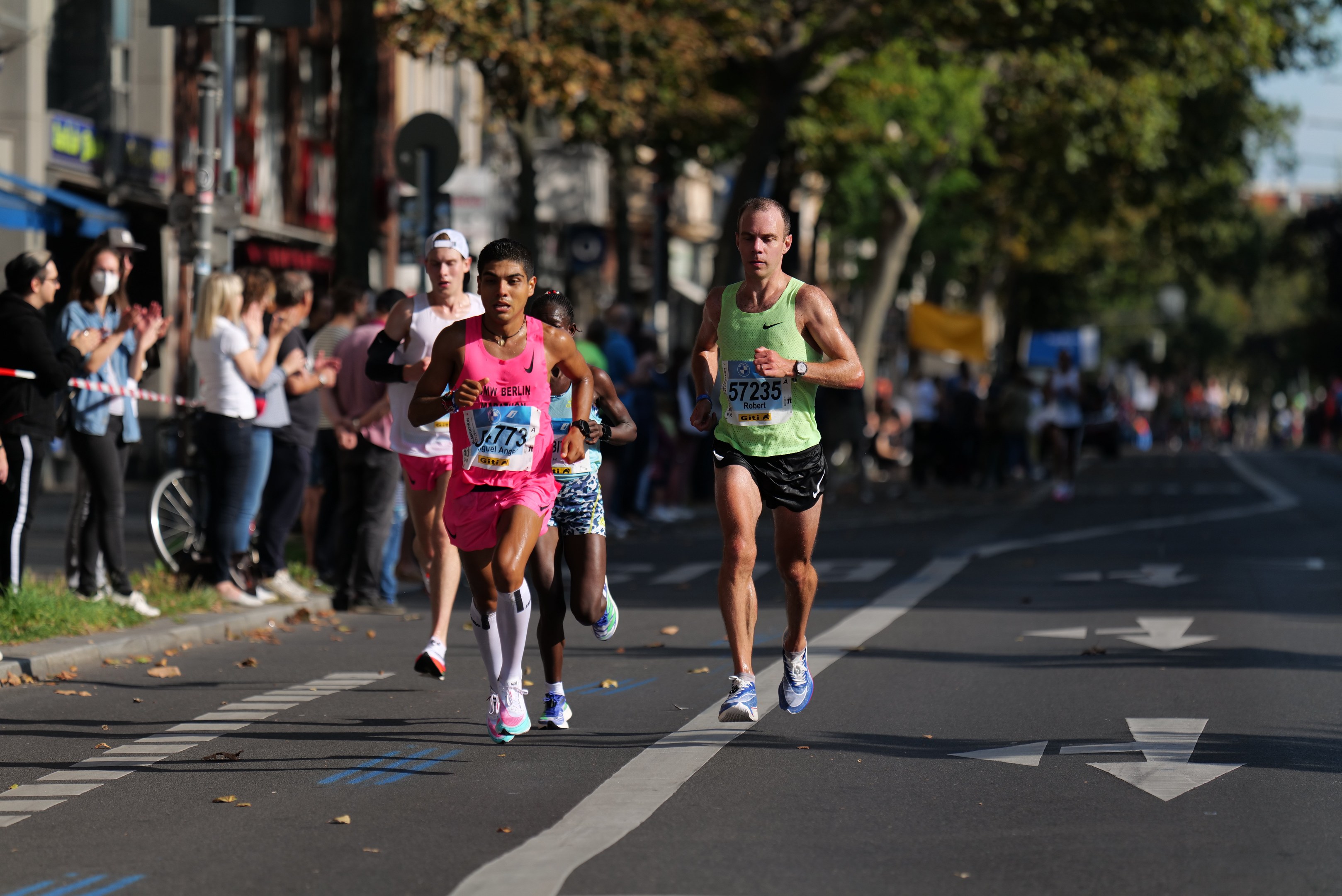Gruppe von Menschen beim Marathon auf einer Stadtstraße mit Zuschauern, Bäumen, Gebäuden und einem Fahrrad im unscharfen Hintergrund.