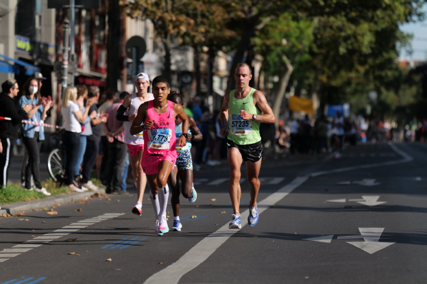 Gruppe von Menschen beim Marathon auf einer Stadtstraße mit Zuschauern, Bäumen, Gebäuden und einem Fahrrad im unscharfen Hintergrund.