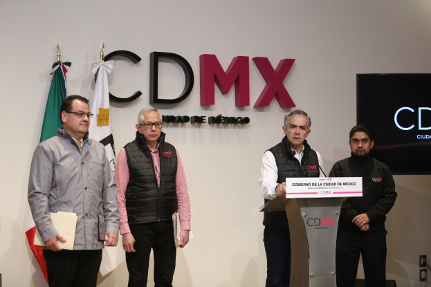 Vier Männer stehen vor einem Podium mit einem Mikrofon und einer Tafel mit der Aufschrift "CDMX, Mexikos erste digitale Währungsbörse"; zwei halten Dateien, zwei tragen Brillen, mit Flaggen und einem Bildschirm im Hintergrund.