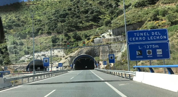 Auto fährt durch einen Tunnel auf einer Straße mit Geländern, Schildern, Polen, Verkehrssignalen, Bäumen, Felsen und einem klaren blauen Himmel.
