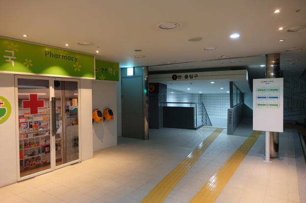 Apothekeneingang mit einem 'Apotheke'-Schild auf der linken Seite, einer Tafel mit Text auf der rechten Seite, beleuchteten Lampen darüber und Hintergrundobjekten.