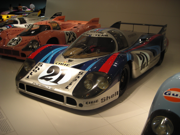 Ein Porsche 917K Rennwagen auf der Le Mans in einem Museum ausgestellt, umgeben von anderen Sportwagen und Menschen, mit einer Wand und einem informativen Schild im Hintergrund.