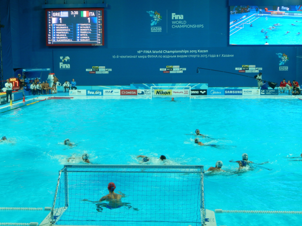 Wasserballspieler bei den 2016 FINA-Weltmeisterschaften in Kazan im Pool gegeneinander antretend, während Zuschauer links sitzen und ein Scoreboard mit Event-Branding im Hintergrund angezeigt wird.