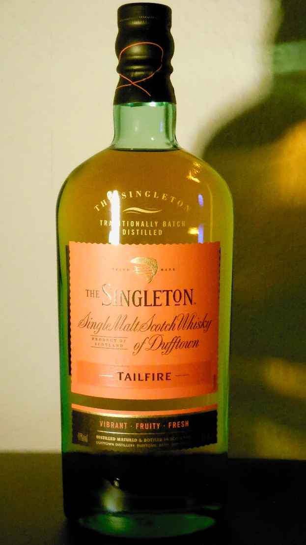 Eine tiefe bernsteinfarbene Flasche Singleton of Dufftown Tailfire Single Malt Scotch Whisky auf einem Tisch, mit einem schwarzen Etikett, das eine Löwen- und Burgwappen zeigt.
