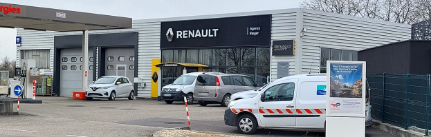 Ein Renault-Autohaus mit parkenden Autos davor, einem Metallzaun, einer Texttafel, Pfosten, einem Schild, Bäumen und einem bewölkten Himmel.