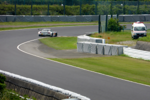 Ein Rennwagen fährt eine Rennstrecke hinunter, im Hintergrund ein Polizeiauto, umgeben von Grünfläche und Absperrung, mit Text unten auf dem Bild.
