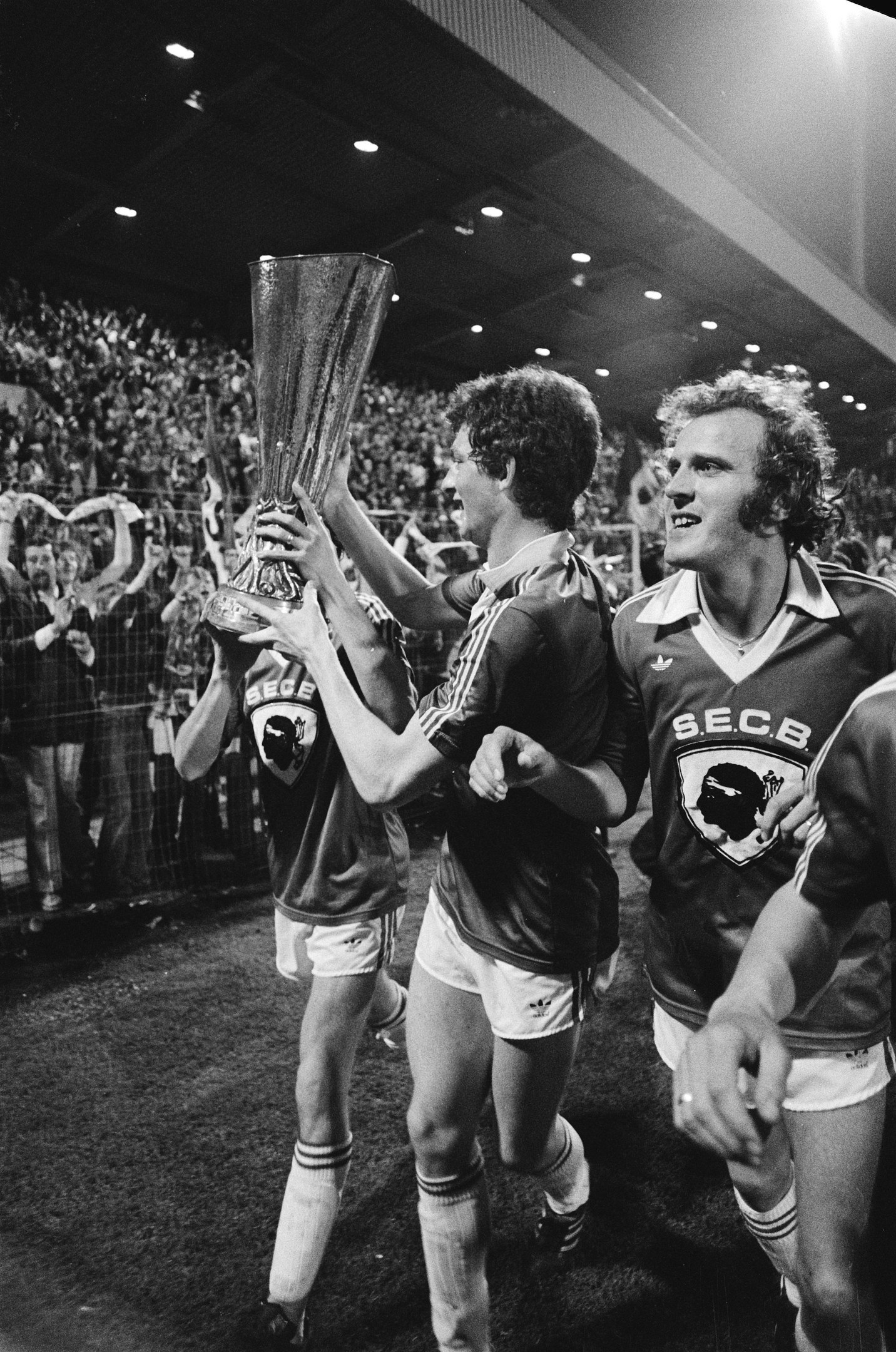 Schwarzes und weißes Bild von Fußballspielern, die auf einem Feld einen Pokal hochhalten, mit einer Menge im Hintergrund.