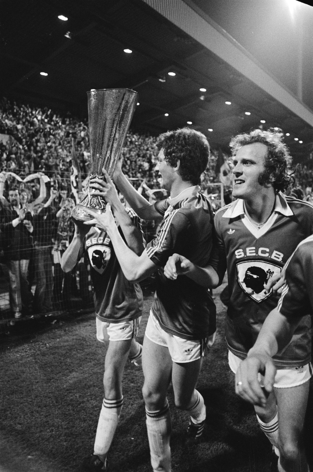 Schwarzes und weißes Bild von Fußballspielern, die auf einem Feld einen Pokal hochhalten, mit einer Menge im Hintergrund.