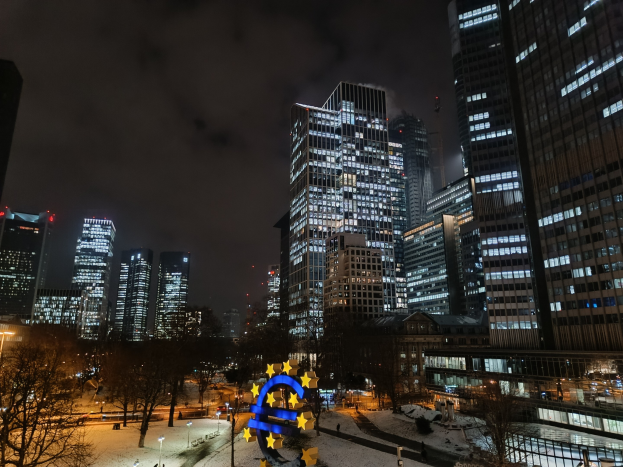 Europäischer Zentralbank-Hauptsitz in Frankfurt, Deutschland, nachts mit beleuchteten Gebäuden, Bäumen, Straßenlaternen, Lichtern und Fußgängern unter einem bewölkten Himmel.