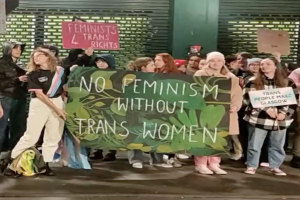 Gruppe von Menschen mit einem Banner, auf dem "Kein Feminismus ohne Transfrauen" steht, und Schildern in einem öffentlichen Raum mit einer Wand und einem Rollladen im Hintergrund.