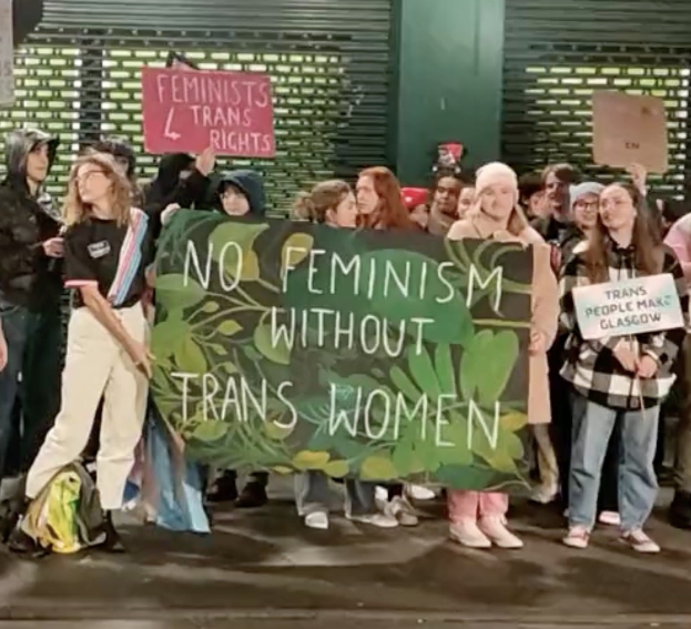 Gruppe von Menschen mit einem Banner, auf dem "Kein Feminismus ohne Transfrauen" steht, und Schildern in einem öffentlichen Raum mit einer Wand und einem Rollladen im Hintergrund.