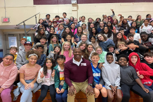 Gruppe von Schülern, die in einem Turnsaal für ein Foto posieren, einige sitzen auf der Treppe und andere stehen, eine Tür und ein Ausgangsschild auf der linken Seite sichtbar, was auf ein Sommerlager hindeutet.