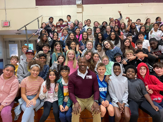 Gruppe von Schülern, die in einem Turnsaal für ein Foto posieren, einige sitzen auf der Treppe und andere stehen, eine Tür und ein Ausgangsschild auf der linken Seite sichtbar, was auf ein Sommerlager hindeutet.