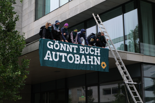 Gruppe von Menschen auf einem Gebäudedach mit einer Fahne, einem Baum links und Glasfenstern im Hintergrund, die zu protestieren scheinen.