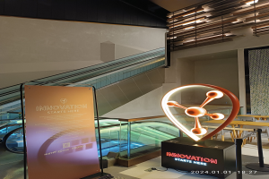 Ein großes Schild in einem Gebäude mit einer Rolltreppe im Hintergrund, Tischen und Stühlen auf der rechten Seite und einer Tafel mit der Aufschrift "Innovation beginnt hier" auf der linken Seite, die auf die HSTP20 Expo hinweist.