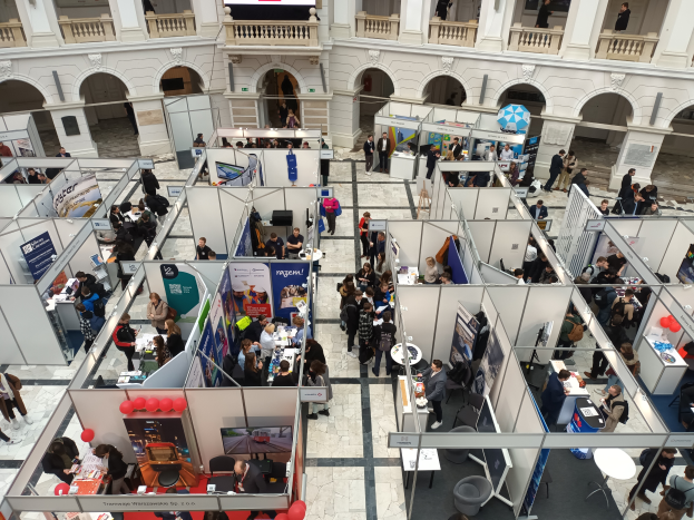 Eine große Gruppe von Menschen versammelte sich in einem Raum voller Stände, Banner, Tische, Stühle und Luftballons, mit einem Gebäude mit Bögen, Säulen, Geländern und einem Bildschirm im Hintergrund.